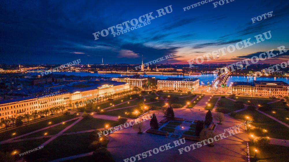 На фотографии изображен ночной Петербург с высоты. Дорожные фонари и освещение зданий рисуют тёплые линии света, а мост через Неву дополняет городской пейзаж. Фотография может использоваться в качестве иллюстративного материала в туристических брошюрах, для украшения интерьеров или в образовательных целях, чтобы показать красоту ночного города.